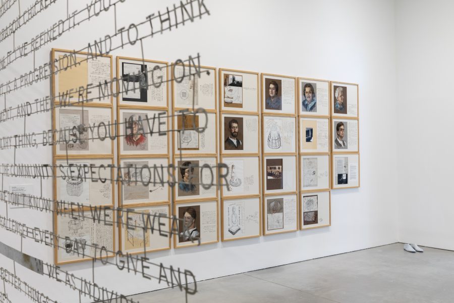 Art exhibition featuring framed sketches, notes, and portraits on a wall. Suspended metal text partially obstructs the view. The floor is smooth concrete, and a pair of cast white shoes is placed in the corner.